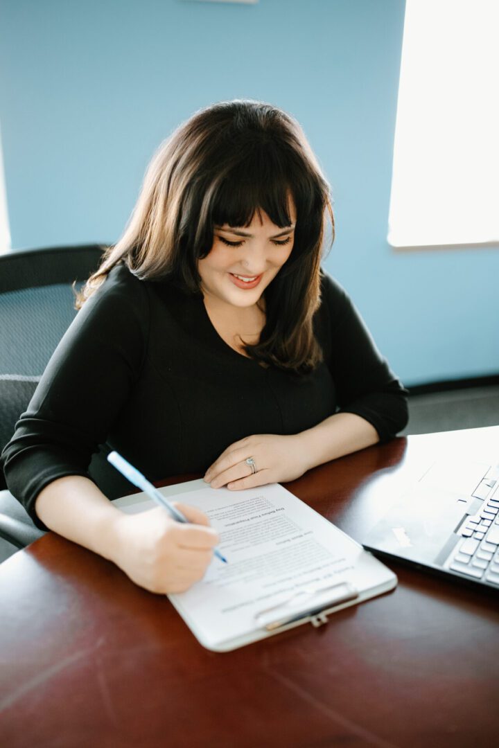 A woman happily writing notes at her desk with a laptop nearby.