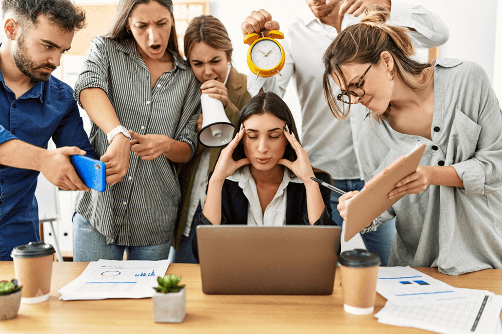 Stressed woman at laptop surrounded by busy colleagues with a ringing alarm clock.